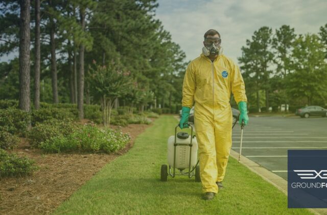 A licensed herbicide applicator in protective gear treating a large commercial turf area with a backpack sprayer.